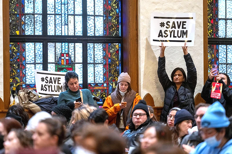 Woman inside a large room with a crowd of people gathered. Woman is holding sign above her head that says #SaveAsylum with a serious expression.