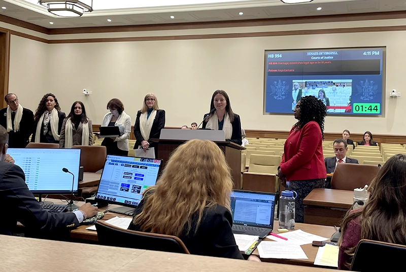 People gathered in a government presentation room with, some wearing white scarves in solidarity. A white woman with long brown hair is presenting at the podium.