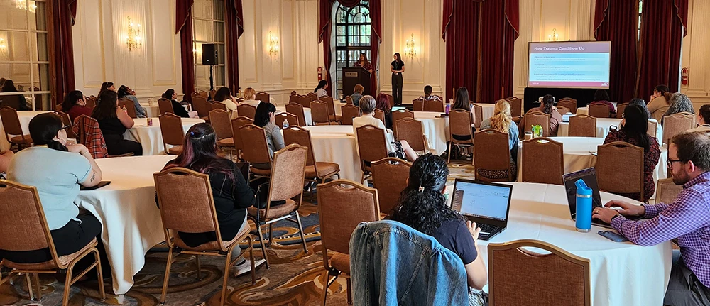 Two women standing in front of a presentation screen with title "The Unique and Complex Challenges of Creating Safety Alongside Immigrant Survivors of Sexual Assault"