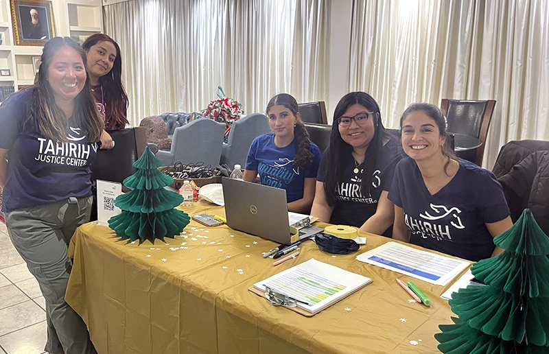 Group of smiling young women wearing Tahirih justice center tshirts and sitting behind a table with gold tablecloth