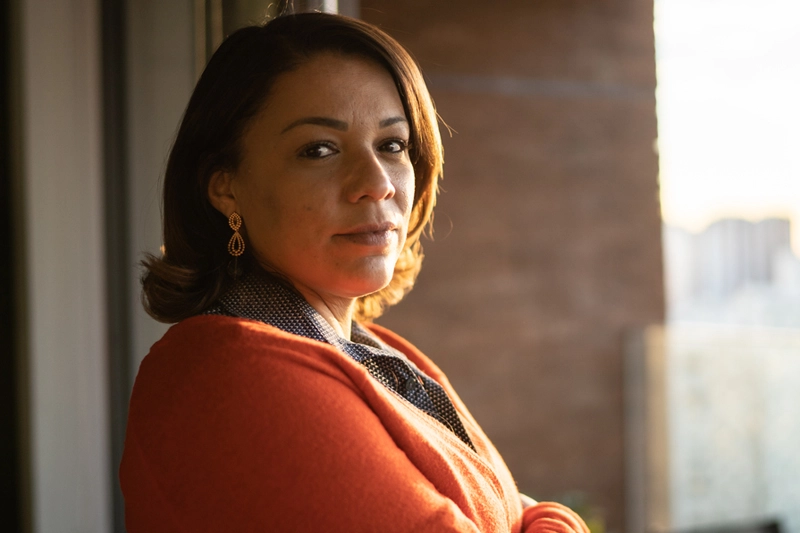 Middle aged Latina woman with serious expression standing by a window and looking back at the camera.